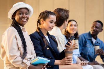 Friends with disposable coffee cup talking in college park. International group of students young men and women enjoying break at university campus, using gadgets, have conversation