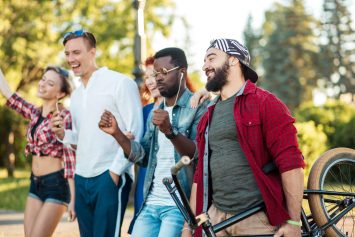Multicultural group of friends with headphones walking together holding bmx bike in the public park - Interracial happy couples having fun day outdoor.